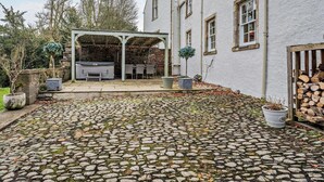 A stone-paved courtyard with a covered outdoor seating area, a hot tub, and a white building with multiple windows. - 4 Bed in Hawick (oc-95765) (Teviotdale)