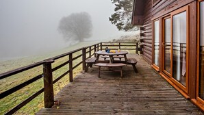 Une terrasse en bois avec une table de pique-nique et des chaises, donnant sur un paysage brumeux avec un arbre.