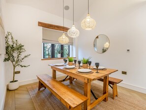 Un coin repas avec une table en bois, des bancs, des luminaires suspendus en osier, une plante en pot et une fenêtre donnant sur des arbres.