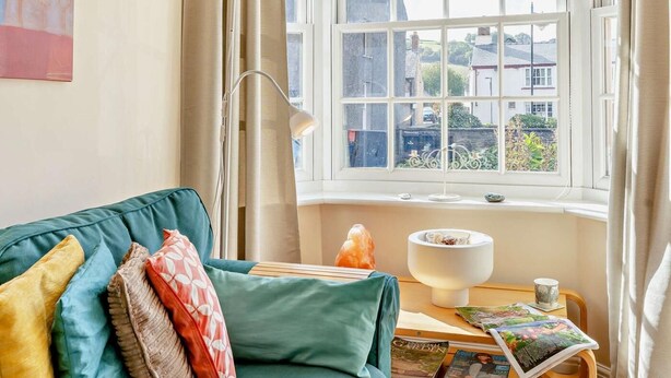 Ein gemütliches Zimmer mit einem türkisfarbenen Sofa, einer Lampe, einem Fenster mit Blick auf Gebäude und einem Tisch mit Zeitschriften.