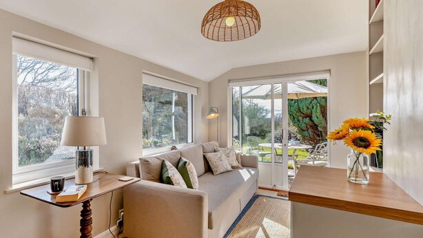 A modern living room with a sofa, a table with a lamp, a bookshelf, and a view of the garden through large windows. - 2 Bed in Grange-over-sands (oc-95811) (Crosthwaite and Lyth)