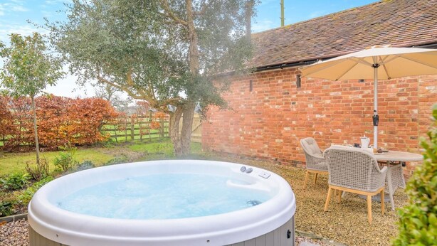 A hot tub in a backyard with a brick wall, a wooden deck, and a table with chairs. - 1 Bed in Gloucester (oc-95793) (Tirley)