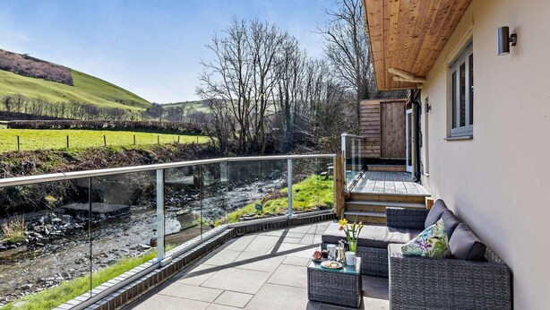 A modern outdoor patio with a glass balustrade overlooking a stream, a seating area with cushions, and a wooden deck. - 2 Bed in Llanidloes (oc-95815) (Llanidloes Without)
