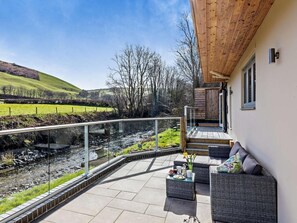 Eine moderne Außenpatio mit Glasbrüstung, Blick auf einen Bach, eine Sitzgruppe mit Kissen und ein Holzterrasse.