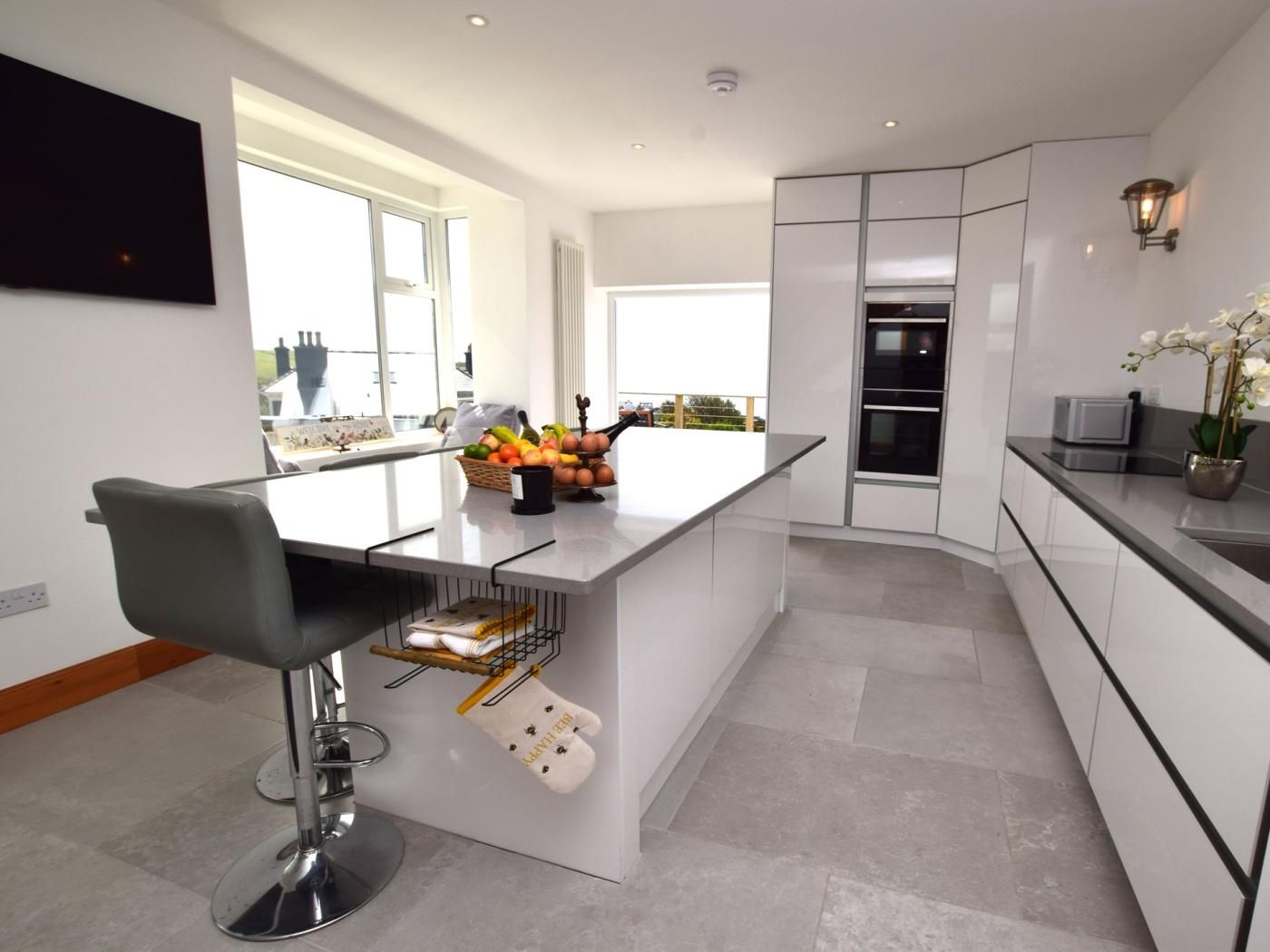 A modern kitchen with a central island, bar stools, a fruit basket, and a view of the outside.