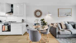 Una cocina moderna con armarios blancos, un comedor con mesa de vidrio y sillas grises, y un sofá gris con un tapete estampado.