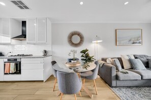 A modern kitchen with white cabinets, a dining area with a glass table and grey chairs, and a grey sofa with a patterned rug. - Two bedroom  in Australia, Henley Beach South (Henley Beach South)