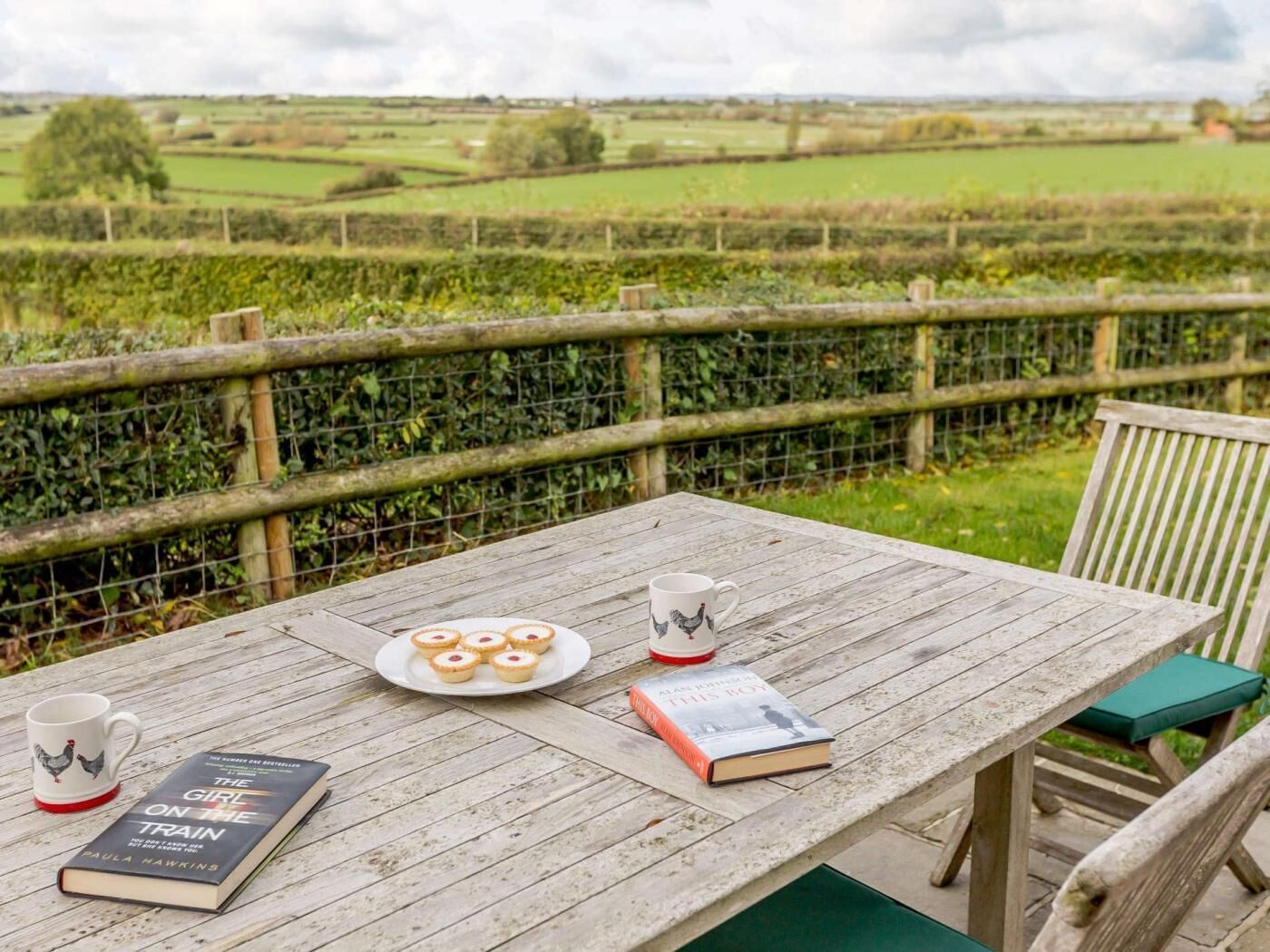 A wooden table with a book, a plate of pastries, and a mug, set against a rural backdrop.