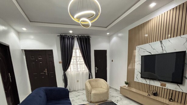 Uma sala de estar moderna com um sofá seccional, uma poltrona bege, uma TV fixada em uma parede de mármore e um lustre.