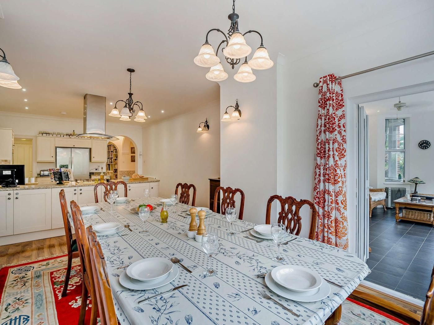 A dining area with a long table set for four, surrounded by chairs, with a kitchen in the background.