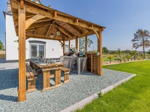 Una pèrgola de fusta a l'aire lliure amb zona de barbacoa, incloent una barbacoa i un safareig, sobre un sòl de grava i amb un jardí al fons.