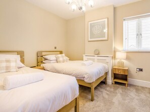 A bedroom with two beds, a chandelier, a framed artwork, a bedside table with a lamp, and a window with blinds.