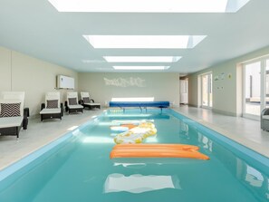 A swimming pool with lounge chairs and a bench, large windows, and a ceiling with skylights.