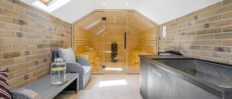 A modern interior with a glass-walled sauna, a seating area with a sofa and cushions, and a large, dark-colored hot tub.