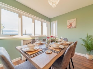 Comedor con mesa de madera dispuesta para cuatro, ventanas amplias y una araña.