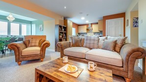 A living room with a sofa, two armchairs, a coffee table with croissants, and a kitchen in the background. - 2 Bed in Bonnybridge (oc-95833) (Bonnybridge)