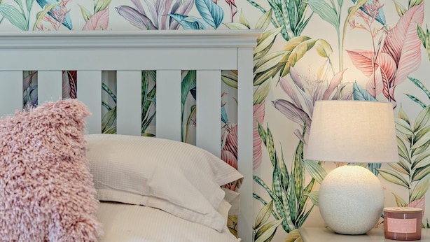 Un lit avec un oreiller rose et moelleux, une tête de lit blanche et une table de chevet sur laquelle se trouvent une lampe et une bougie.