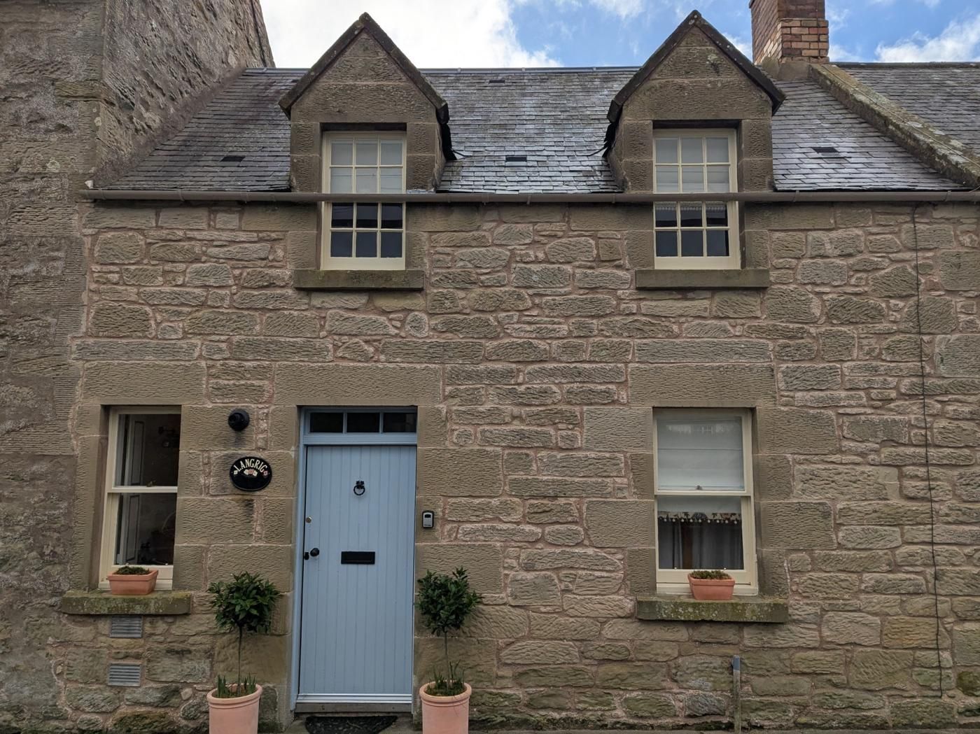 A stone building with a blue door, two windows, and a small porch with potted plants.