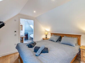 Ett sovrum med ett sluttande tak, en säng med grå sängkläder, ett sängbord med en lampa och utsikt över ett badrum och ett kök genom en öppen dörröppning.