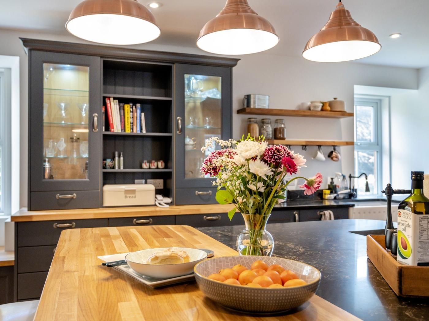 Una cocina con encimera de madera, un bol de naranjas, un jarrón con flores y un panera.