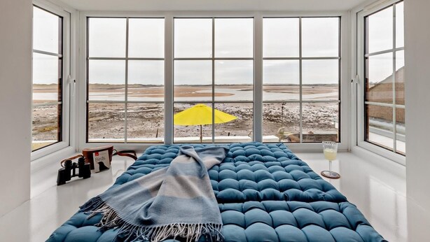 Ein Zimmer mit einem großen Bett, einem gelben Regenschirm und Blick auf eine karge Landschaft durch große Fenster.