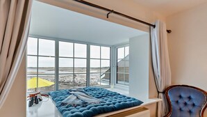 Ein Schlafzimmer mit großem Fenster, einem Bett mit blauem Polsterkopfteil, einem Sessel und Blick auf einen Strand.
