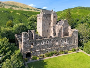 A historic stone castle with multiple towers, surrounded by lush green hills and a well-maintained lawn.