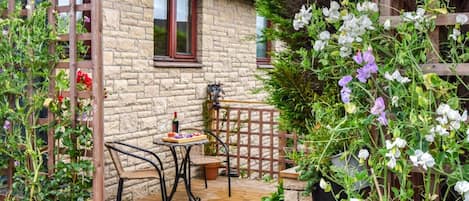A stone house with a wooden deck, a table with a bottle and glass, and a variety of flowering plants.