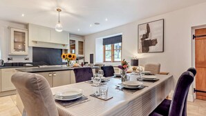 A modern kitchen with white cabinets, a central island, and a dining area with a striped tablecloth and purple chairs. - 4 Bed in Ilfracombe (oc-95899) (Berrynarbor)