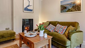 A living room with a green sofa, a wooden coffee table with breakfast items, a fireplace, and two framed pictures on the wall. - 2 Bed in Fowey (oc-91015) (Polruan)