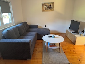 A living room with a grey sectional sofa, a white coffee table, a painting on the wall, and a TV on a wooden stand.