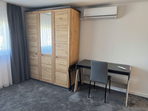 A room with a wooden wardrobe, a desk with two chairs, and an air conditioning unit on the wall.