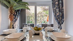 A dining table set with plates, glasses, and a bowl of fruit, with a potted plant and curtains. - 3 Bed in Llandudno (oc-95068) (Llandudno)