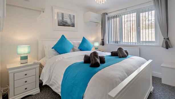 A hotel room with a bed, a nightstand, a lamp, a window with curtains, and a chandelier. - 3 Bed in Llandudno (oc-95068) (Llandudno)