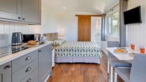 Een compacte kamer met kitchenette, een bed met beddengoed met een patroon, een eettafel met een stoel, en een televisie aan de muur.