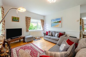 A living room with a flat-screen TV, grey sofa, red and white cushions, a patterned rug, and a painting of a beach scene. - 2 Bed in Honiton (oc-52420) (Honiton)