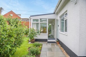 A house with a garden, a conservatory, and a red brick building in the background. - 2 Bed in Honiton (oc-52420) (Honiton)