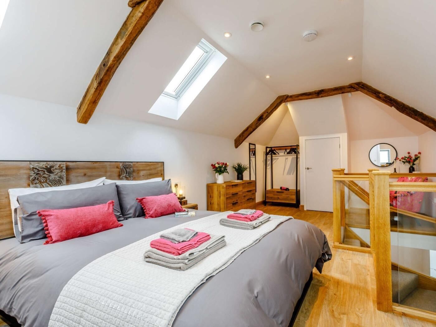 Ein Schlafzimmer mit schrägverdeckter Decke, Holzbalken, einem Oberlicht, einem Bett mit Kissen, einem Nachttisch mit Lampe, einem Schminktisch und einer Treppe.