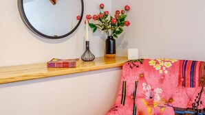 A room with a round mirror, a vase with red flowers, a wooden shelf with books, and a colorful sofa. - 1 Bed in Umberleigh (oc-82998) (Atherington)