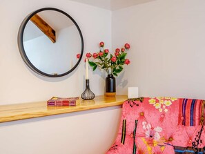 A room with a round mirror, a vase with red flowers, a wooden shelf with books, and a colorful sofa. - 1 Bed in Umberleigh (oc-82998) (Atherington)