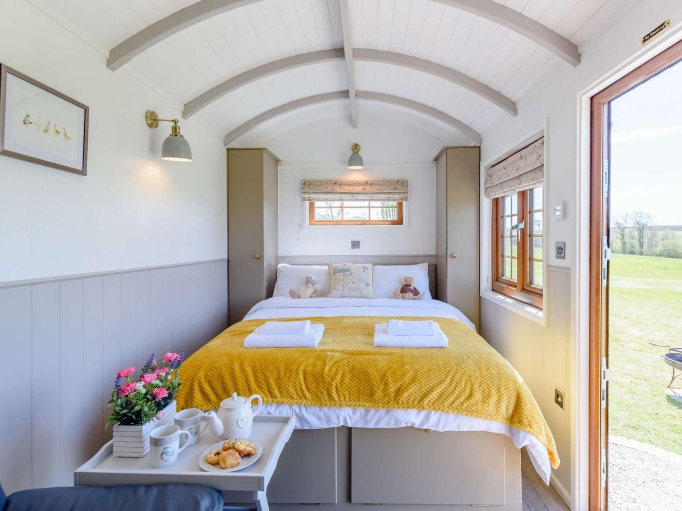 A bedroom with a bed, bedside table with a tray of breakfast items, a window with a view of the outdoors, and a wall-mounted light fixture.