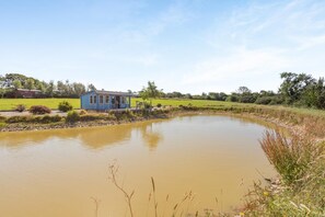 A small blue building by a pond with a grassy field and trees in the background. - 1 Bed in Beaworthy (oc-93267) (Ashwater)