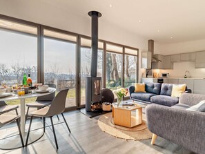 A modern living room with a large window, a fireplace, a dining area with breakfast setup, and a kitchen with contemporary design.
