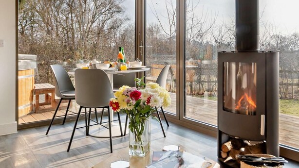 A modern living room with a large window, a fireplace, a dining table with chairs, and a vase with flowers. - 2 Bed in Colchester (oc-95941) (East Bergholt)