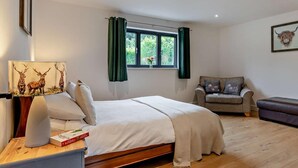 A bedroom with a bed, a bedside table with a lamp and book, a window with curtains, a sofa, and a framed picture on the wall. - 5 Bed in Dulverton (oc-87003) (Morebath)