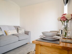 A modern living room with a grey sofa, a wooden coffee table, and a dining area with white plates and a vase of flowers. - 1 Bed in Okehampton (oc-78799) (Hatherleigh)