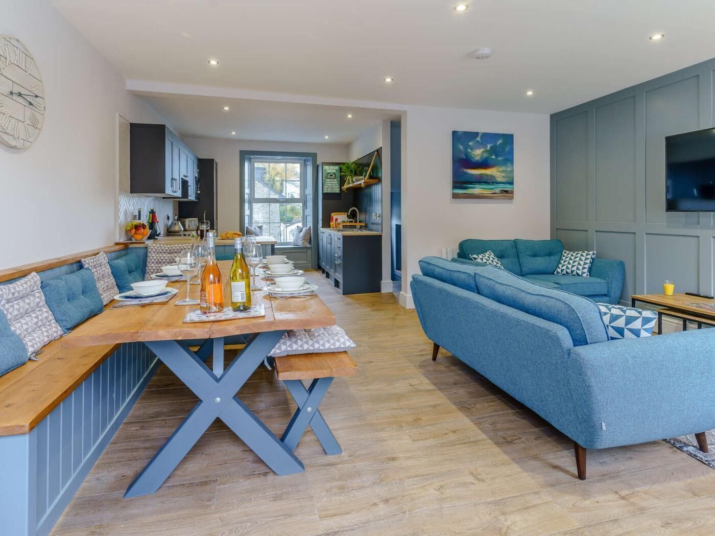 A modern living room with a blue sofa, a wooden dining table set for a meal, and a kitchen in the background.