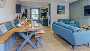 A modern living room with a blue sofa, a wooden dining table set for a meal, and a kitchen in the background. - 3 Bed in New Quay (oc-83998) (New Quay)