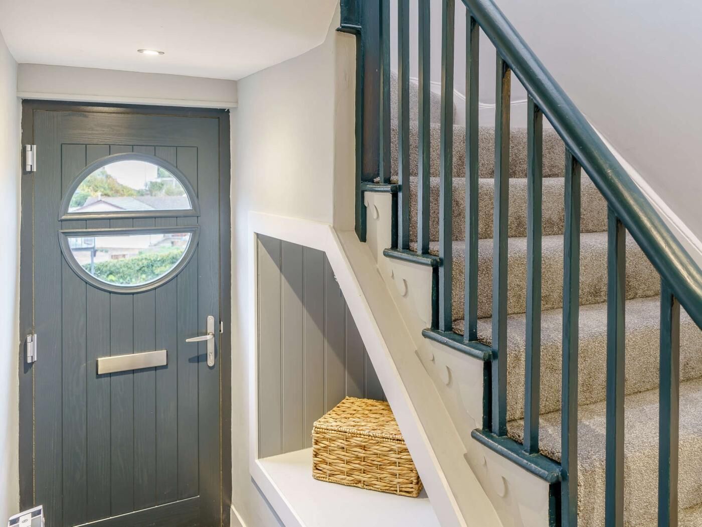 A dark wooden door with a window, a white wall, and a staircase with beige carpet and black railings.