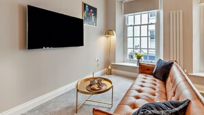 Ein Wohnzimmer mit einem Ledersofa, einem Couchtisch, einem an der Wand montierten Fernseher, einer Stehlampe und einem Fenster mit Blick auf Gebäude.